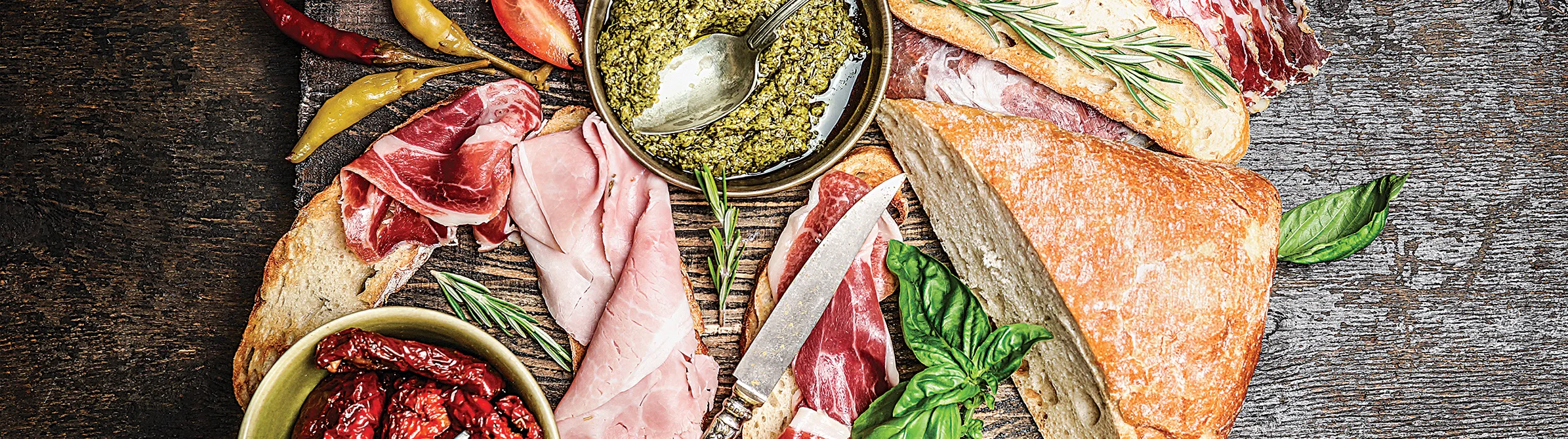 A rustic antipasti spread with bread, cured meats, pesto, peppers, and herbs on a wooden surface.