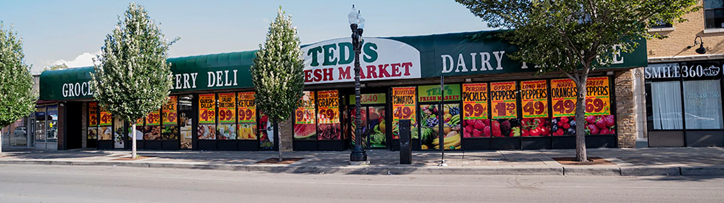 Store front of Ted's Fresh Market with promotional posters on the windows and trees lining the sidewalk